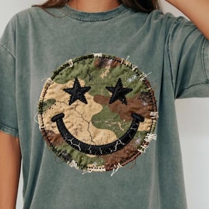 May include: A faded green t-shirt with a large, circular patch displaying a camouflage pattern and a smiley face design. The smiley face has black star eyes and a curved black mouth. The patch is stitched onto the shirt with visible thread.