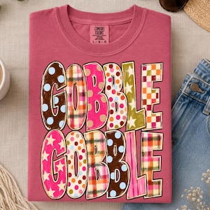 May include: A dusty rose crewneck t-shirt featuring the text GOBBLE GOBBLE in large, multi-patterned letters. The letters display various designs including polka dots, plaid, and stars in colors like brown, pink, orange, yellow, and blue.