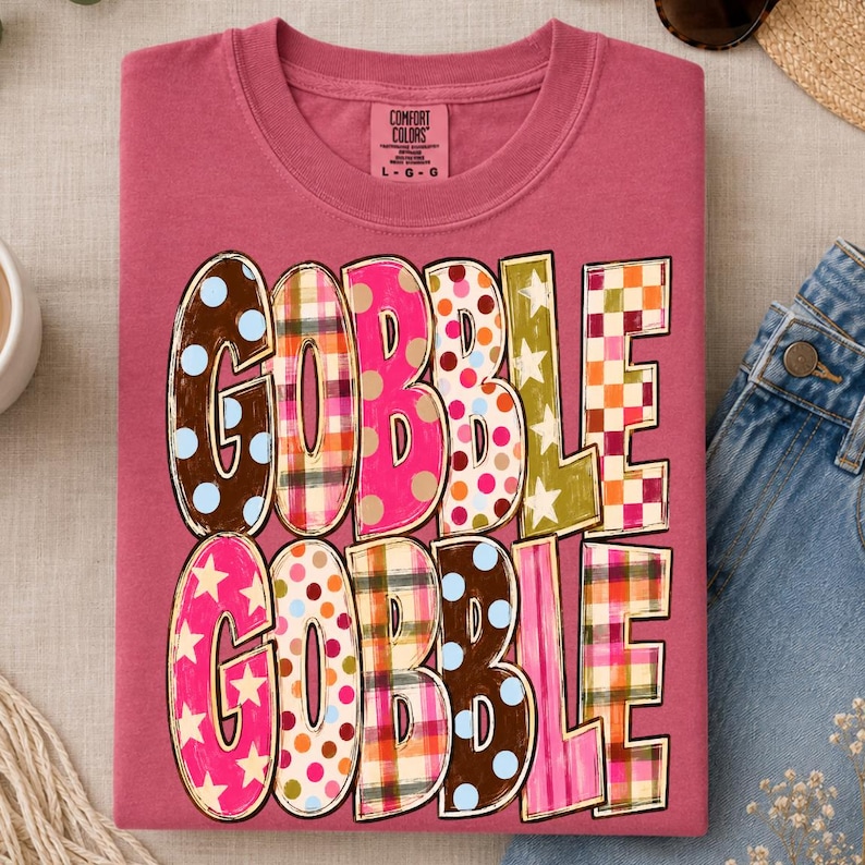 May include: A dusty rose crewneck t-shirt featuring the text GOBBLE GOBBLE in large, multi-patterned letters. The letters display various designs including polka dots, plaid, and stars in colors like brown, pink, orange, yellow, and blue.