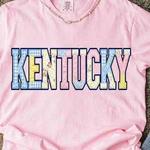 May include: A light pink t-shirt with the word "KENTUCKY" in a patchwork design across the front. The letters are outlined in navy blue and filled with various patterns, including gingham, floral, and polka dots. A delicate silver chain necklace is around the neck.