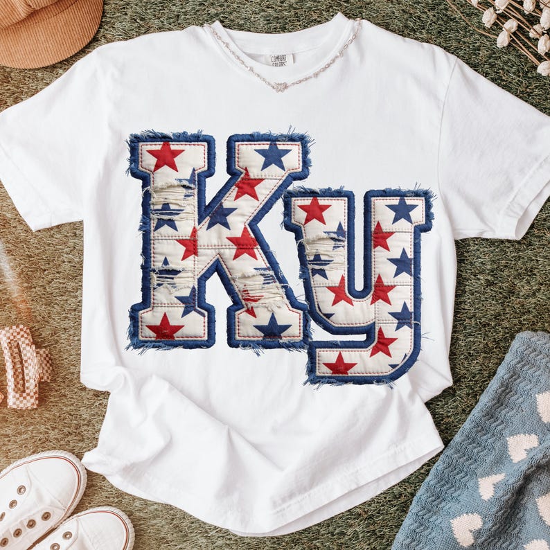 May include: White t-shirt featuring the letters "KY" in a distressed design. The letters are filled with red and blue stars, and outlined with a frayed denim-like trim. The shirt is a casual, comfortable style.
