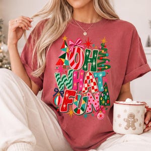 K&ouml;nnte beinhalten: Ein ziegelrotes Kurzarm-T-Shirt mit einem bunten Grafikdruck, der den Text "OH WHAT FUN" in verschiedenen Mustern und Farben zeigt, umgeben von Illustrationen von Weihnachtsb&auml;umen, Ornamenten, Sternen und einer rosa Schleife.