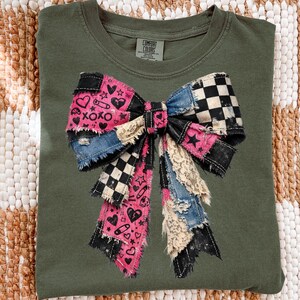 Op de afbeelding: Olijfgroen T-shirt met een groot patchwork strik ontwerp. De strik heeft denim, zwart-wit geruite, roze en kanten stoffen. De roze secties hebben harten, sterren en de tekst "XOXO". Het shirt heeft een Comfort Colors label.