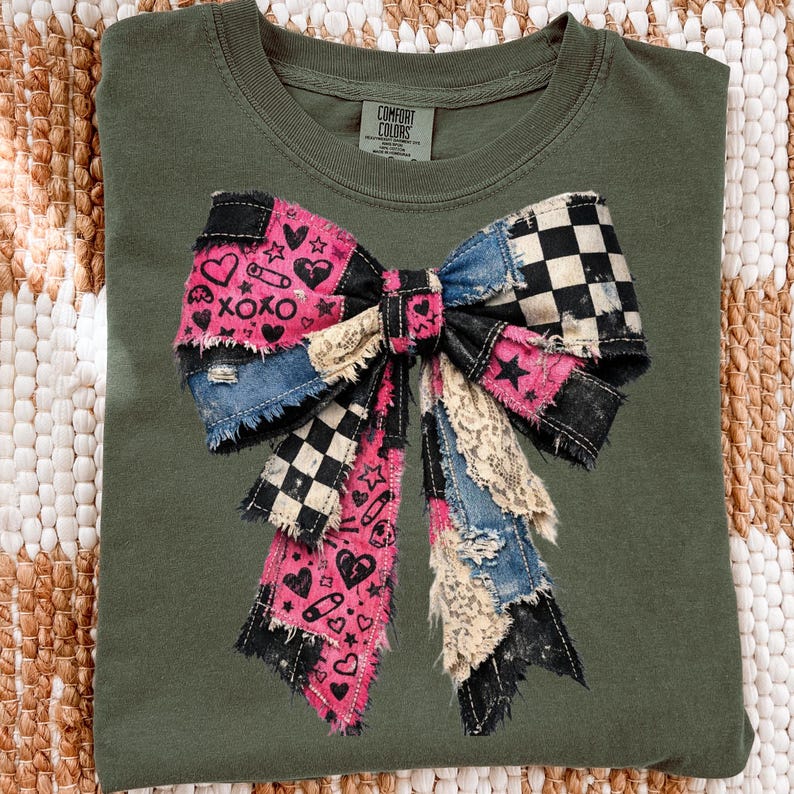 Op de afbeelding: Olijfgroen T-shirt met een groot patchwork strik ontwerp. De strik heeft denim, zwart-wit geruite, roze en kanten stoffen. De roze secties hebben harten, sterren en de tekst "XOXO". Het shirt heeft een Comfort Colors label.