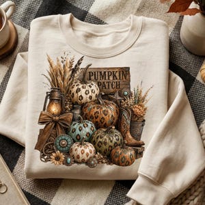 May include: Cream-colored sweatshirt featuring a "Pumpkin Patch" design. The graphic includes various pumpkins with leopard print and other patterns, a lantern, cowboy boots, and decorative elements. The design is set against a rustic wooden sign.