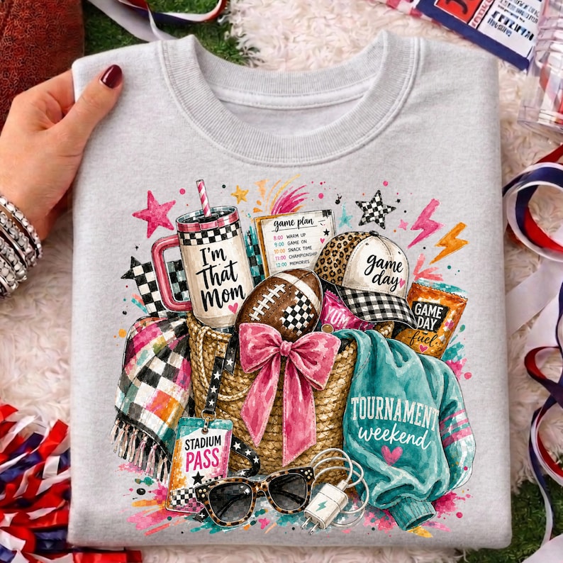 May include: A light gray sweatshirt featuring a graphic print of a straw basket filled with a tumbler labeled "I'm That Mom," a football, a baseball cap labeled "game day," a "stadium pass," sunglasses, and a teal shirt labeled "tournament weekend."