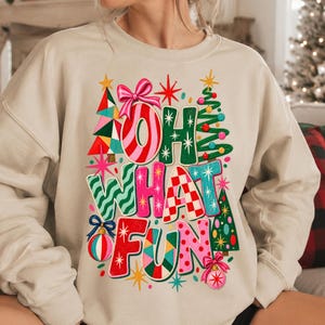 K&ouml;nnte beinhalten: Ein beigefarbenes Sweatshirt mit Rundhalsausschnitt und einem bunten Grafikdruck mit dem Text OH WHAT FUN. Das Design zeigt stilisierte Weihnachtsb&auml;ume, eine rosa Schleife und runde Ornamente in Rot, Gr&uuml;n, Pink und Blau mit verschiedenen Mustern.