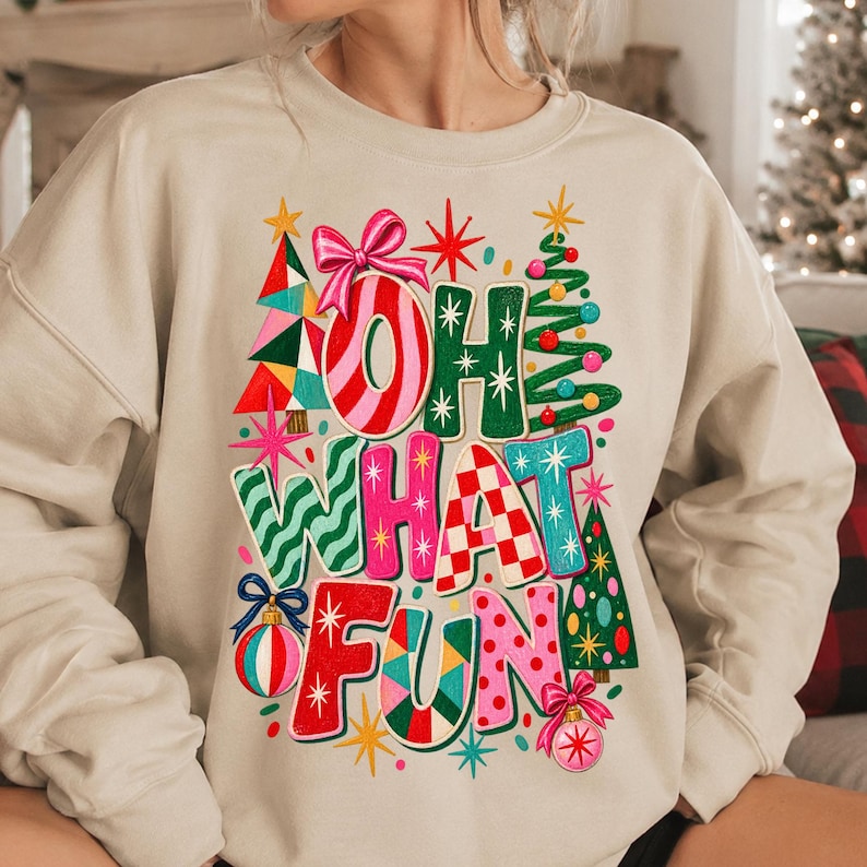 K&ouml;nnte beinhalten: Ein beigefarbenes Sweatshirt mit Rundhalsausschnitt und einem bunten Grafikdruck mit dem Text OH WHAT FUN. Das Design zeigt stilisierte Weihnachtsb&auml;ume, eine rosa Schleife und runde Ornamente in Rot, Gr&uuml;n, Pink und Blau mit verschiedenen Mustern.