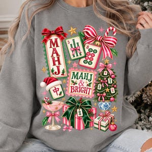 K&ouml;nnte beinhalten: Ein graues Sweatshirt mit Rundhalsausschnitt und einem farbenfrohen, weihnachtlichen Grafikdruck. Das Design zeigt Mahjong-Steine, einen Weihnachtsbaum, Geschenkkartons und ein Cocktailglas sowie den Text "MAHJ & BRIGHT" und "MAHJ" in Rot und Gr&uuml;n.