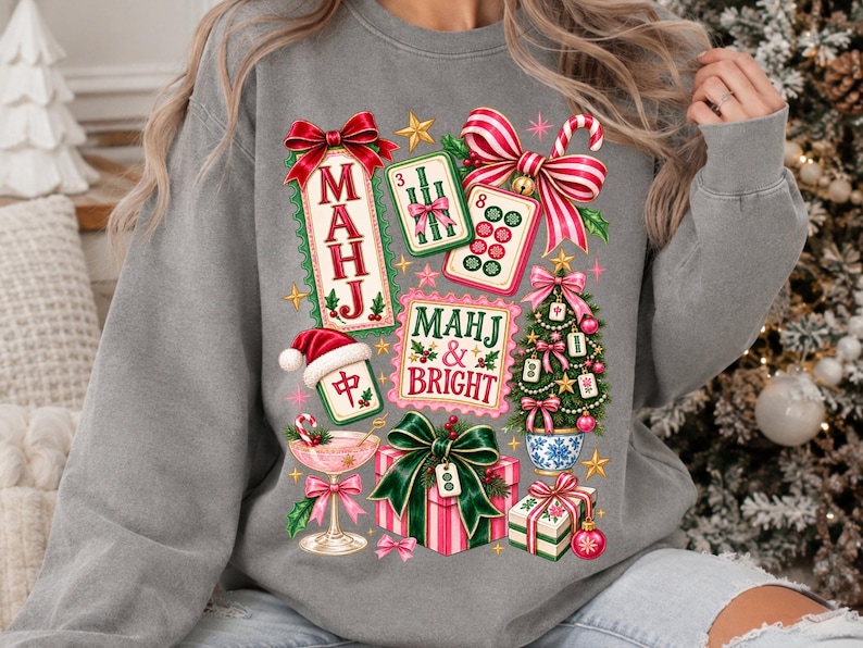 K&ouml;nnte beinhalten: Ein graues Sweatshirt mit Rundhalsausschnitt und einem farbenfrohen, weihnachtlichen Grafikdruck. Das Design zeigt Mahjong-Steine, einen Weihnachtsbaum, Geschenkkartons und ein Cocktailglas sowie den Text "MAHJ & BRIGHT" und "MAHJ" in Rot und Gr&uuml;n.