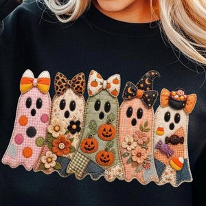 May include: A black sweatshirt featuring a central applique design of five ghosts. Each ghost is decorated with different fabric patterns, bows, and seasonal motifs including pumpkins, flowers, candy corn, and a small cauldron filled with candy.