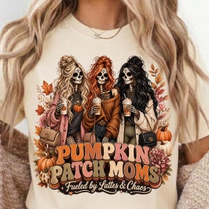 May include: Cream-colored t-shirt featuring a graphic of three skeleton women with long hair, holding coffee cups, and wearing fall-themed outfits. The text "Pumpkin Patch Moms" is displayed below, with the phrase "Fueled by Lattes & Chaos".