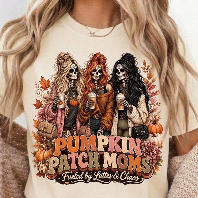 May include: Cream-colored t-shirt featuring a graphic of three skeleton women with long hair, holding coffee cups, and wearing fall-themed outfits. The text "Pumpkin Patch Moms" is displayed below, with the phrase "Fueled by Lattes & Chaos".