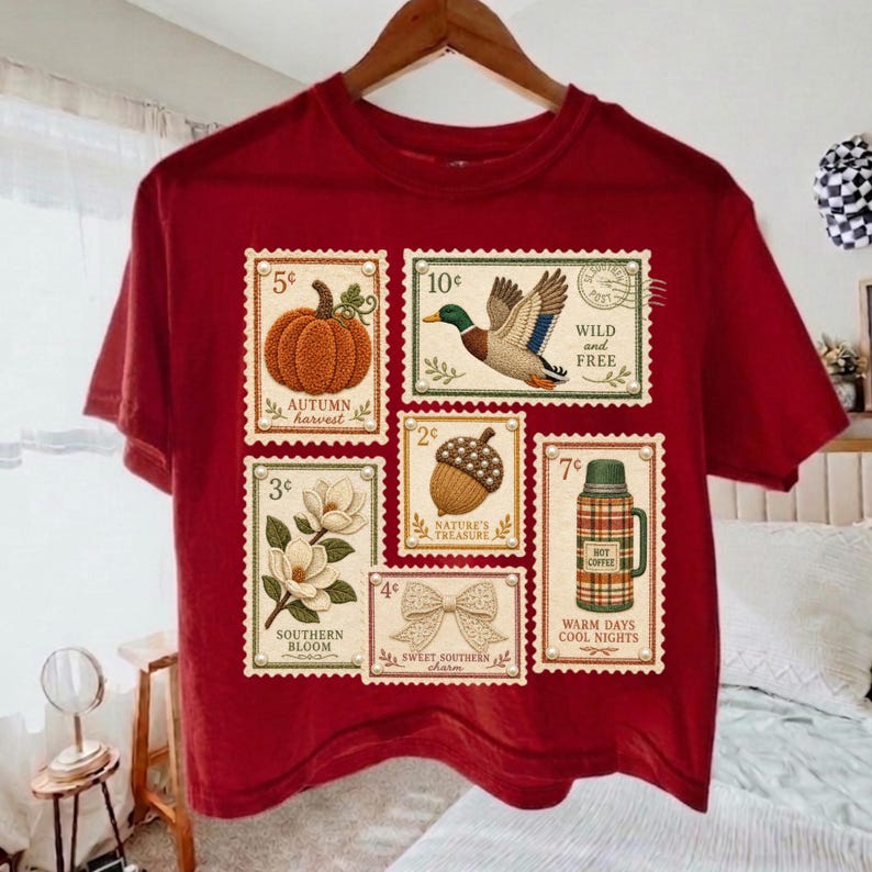 May include: A maroon t-shirt featuring postage stamp designs with autumn and nature themes. Stamps depict a pumpkin, duck, acorn, magnolia, bow, and a thermos. Text includes "Autumn Harvest", "Wild and Free", "Nature's Treasure", "Southern Bloom", "Sweet Southern Charm", "Warm Days Cool Nights".