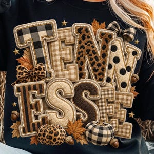 Könnte beinhalten: Ein schwarzes Sweatshirt mit dem Wort TENNESSEE in großen, strukturierten Buchstaben mit verschiedenen Mustern. Die Buchstaben zeigen Karomuster, Leopardenmuster und Punkte, verziert mit kleinen Schleifen, Herbstblättern, Eicheln und goldenen Sternen.