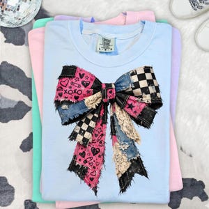 Op de afbeelding: Een lichtblauw Comfort Colors T-shirt met een grote strik-afbeelding. De strik heeft een patchwork-ontwerp met roze harten, zwart-wit geruite patronen, denim en kanten details. Het shirt is opgevouwen met andere pastelkleurige shirts.