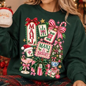 K&ouml;nnte beinhalten: Ein dunkelgr&uuml;nes Sweatshirt mit einem festlichen Grafikdruck aus Mahjong-Steinen, einem Weihnachtsbaum, verpackten Geschenken, einem Cocktailglas und roten Schleifen. Der Text lautet "MAHJ" und "MAHJ & BRIGHT" in dekorativer Schrift, umgeben von goldenen Sternen.