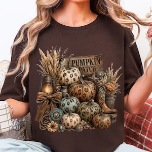 May include: Brown t-shirt featuring a fall-themed graphic. The design includes pumpkins with leopard print, a wooden sign that reads "Pumpkin Patch", and decorative elements like wheat, a lantern, and cowboy boots.