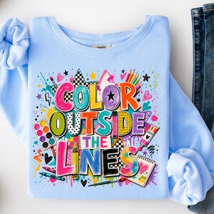 May include: A light blue crewneck sweatshirt featuring a colorful graphic print with the text "COLOR OUTSIDE THE LINES". The design includes illustrations of paint palettes, crayons, paint tubes, sketchbooks, stars, hearts, and paint splatters.