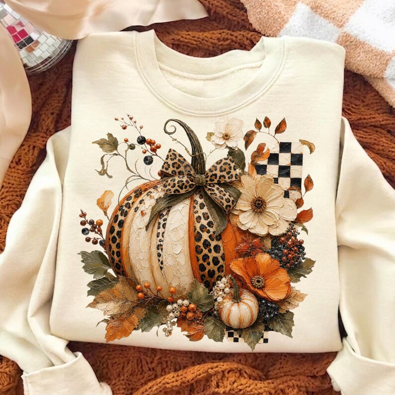 May include: A cream-colored crewneck sweatshirt featuring a central graphic of a large pumpkin with leopard print sections, a leopard print bow, and surrounding autumn foliage, white flowers, orange blossoms, and a black and white checkered pattern.