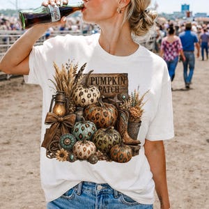 May include: White t-shirt featuring a fall-themed graphic with pumpkins, a lantern, and the text "PUMPKIN PATCH". The design includes various pumpkins, wheat, and a pair of cowboy boots, evoking a rustic autumn aesthetic.