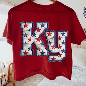 May include: Red t-shirt with the letters "KY" in a distressed denim design. The letters are decorated with red and blue stars, and have a frayed denim border. The shirt is hanging on a wooden hanger.