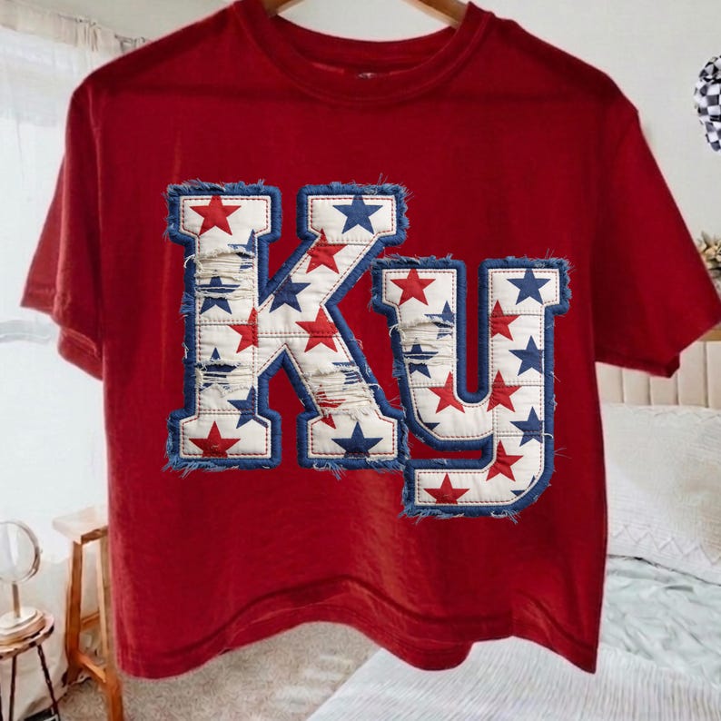 May include: Red t-shirt with the letters "KY" in a distressed denim design. The letters are decorated with red and blue stars, and have a frayed denim border. The shirt is hanging on a wooden hanger.