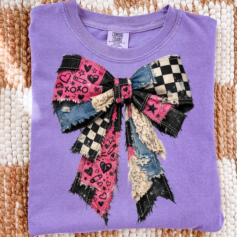 Op de afbeelding: Een paars Comfort Colors T-shirt met een groot patchwork strik ontwerp. De strik bevat denim, zwart-wit geruite en roze stof met harten, sterren en de tekst "XOXO". Het shirt heeft een ronde hals en een label.