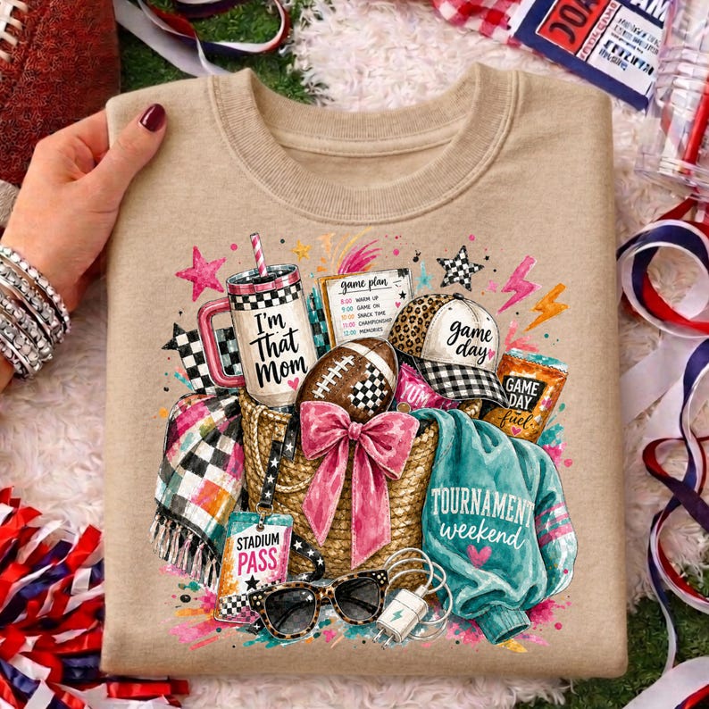 May include: A tan crewneck sweatshirt featuring a graphic print of a straw tote bag filled with a tumbler labeled I'm That Mom, a football, a leopard print cap, a teal jacket, a plaid scarf, sunglasses, and a stadium pass with text.