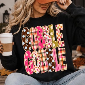 May include: A black long-sleeve sweatshirt featuring the text GOBBLE GOBBLE in large, blocky letters. Each letter displays a different pattern, including brown with blue dots, red and green plaid, pink with stars, and orange and white checkers.