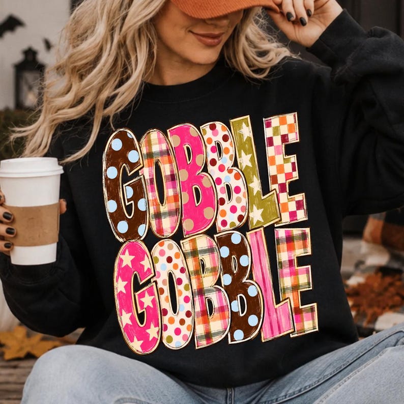 May include: A black long-sleeve sweatshirt featuring the text GOBBLE GOBBLE in large, blocky letters. Each letter displays a different pattern, including brown with blue dots, red and green plaid, pink with stars, and orange and white checkers.