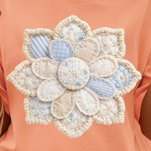 May include: Peach-colored t-shirt featuring a large, appliqued flower. The flower has cream-colored, blue, and beige fabric petals with a crocheted border. The center of the flower is a small circle with a cross-stitch design.