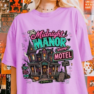 May include: A lilac-colored t-shirt featuring a spooky haunted house design with the text "The Midnight Manor Motel". The graphic includes ghosts, pumpkins, and the words "Vacancy" and "No Refunds".