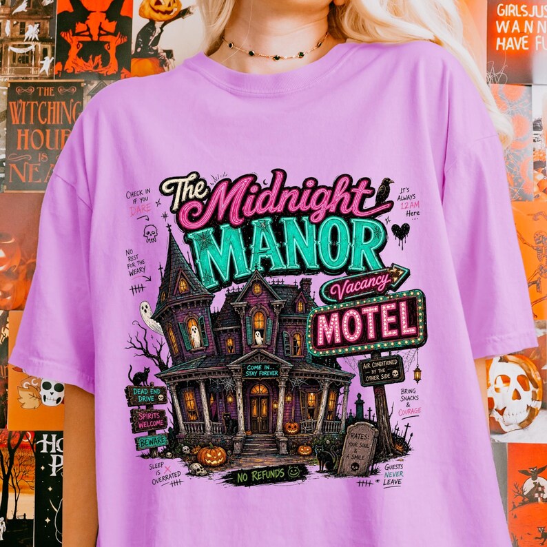 May include: A lilac-colored t-shirt featuring a spooky haunted house design with the text "The Midnight Manor Motel". The graphic includes ghosts, pumpkins, and the words "Vacancy" and "No Refunds".