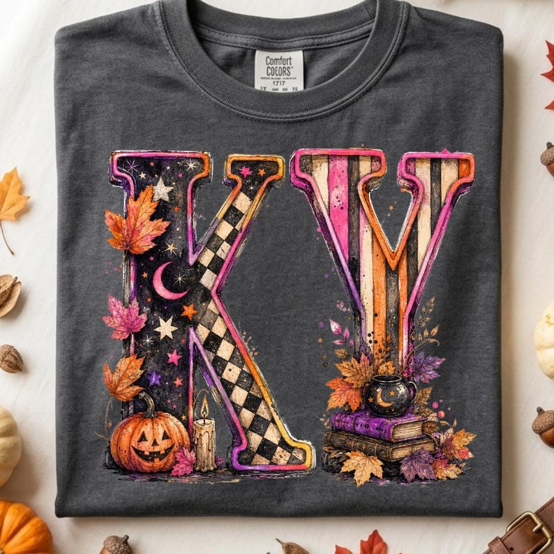 Pode incluir: Uma t-shirt cinzenta escura com um gr&aacute;fico grande das letras KY. O K &eacute; preto com um padr&atilde;o xadrez, estrelas, uma lua crescente, uma ab&oacute;bora e uma vela. O Y tem riscas cor-de-rosa e cor de laranja com livros e um pequeno caldeir&atilde;o.