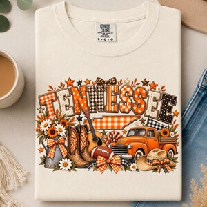 Może przedstawiać: Kremowy t-shirt z nadrukiem graficznym słowa TENNESSEE w pomarańczowe i czarne wzory. Projekt zawiera gitarę, kowbojki, piłkę do futbolu amerykańskiego, pomarańczowy pickup, kowbojski kapelusz oraz słoneczniki ozdobione wstążkami.