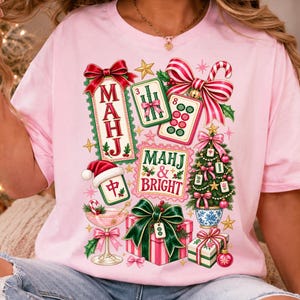 K&ouml;nnte beinhalten: Ein hellrosa T-Shirt mit einem festlichen Aufdruck und dem Text "MAHJ & BRIGHT". Das Design zeigt Mahjong-Steine, einen mit Steinen geschm&uuml;ckten Weihnachtsbaum, Geschenke mit gr&uuml;nen Schleifen, ein Cocktailglas sowie rot-wei&szlig; gestreifte Schleifen.
