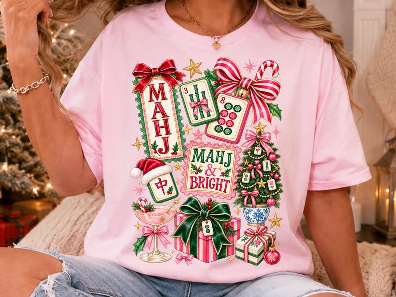 K&ouml;nnte beinhalten: Ein hellrosa T-Shirt mit einem festlichen Aufdruck und dem Text "MAHJ & BRIGHT". Das Design zeigt Mahjong-Steine, einen mit Steinen geschm&uuml;ckten Weihnachtsbaum, Geschenke mit gr&uuml;nen Schleifen, ein Cocktailglas sowie rot-wei&szlig; gestreifte Schleifen.