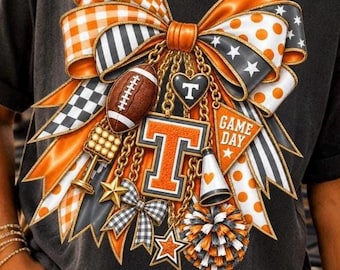 May include: A black t-shirt featuring a large graphic of a decorative bow in orange, white, and black. The bow is adorned with charms including a football, a letter T, a megaphone, stars, and a pennant with the text GAME DAY printed on it.