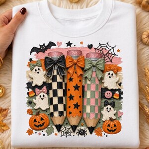 K&ouml;nnte beinhalten: Wei&szlig;es Sweatshirt mit einem Halloween-Motiv. Das Design zeigt drei Bleistifte mit Schleifen, Geistern, K&uuml;rbissen, Flederm&auml;usen und Spinnweben. Die Bleistifte sind schwarz-wei&szlig; kariert, orange mit Sternen und rosa-gr&uuml;n kariert.