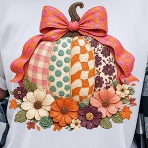 Può includere: Una t-shirt bianca con la grafica di una zucca decorata con pannelli alternati a quadretti rosa, pois verde acqua, strisce ondulate arancioni e motivi floreali viola, sormontata da un fiocco a scacchi rosa e arancione e circondata da fiori.