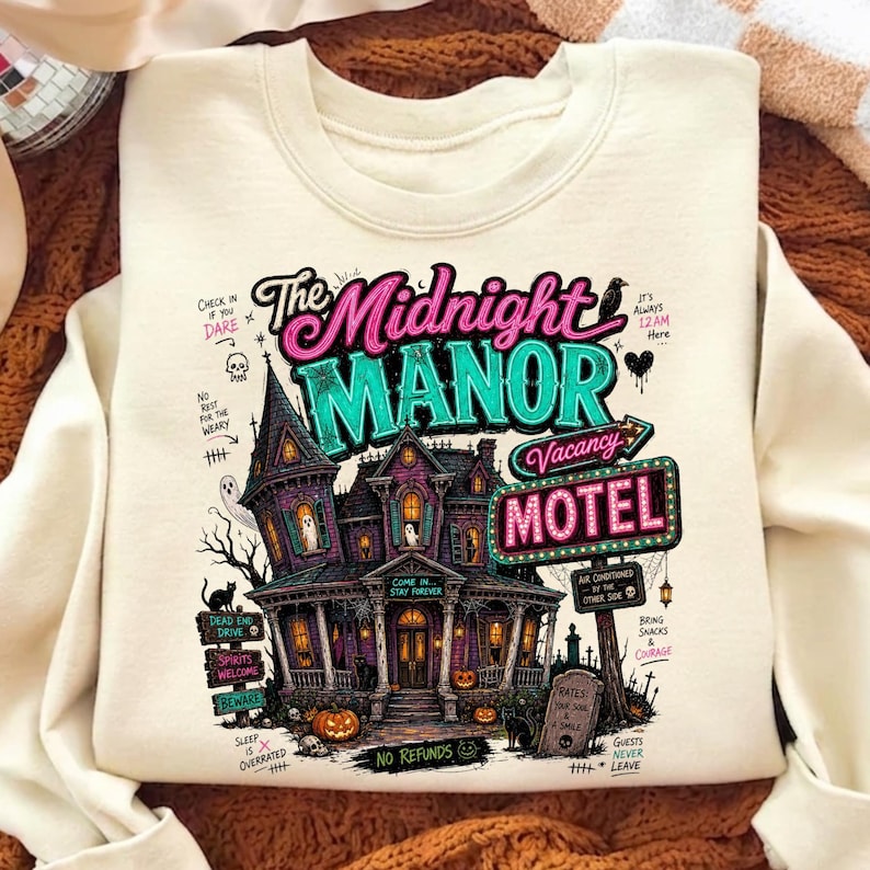 May include: Cream-colored sweatshirt featuring a colorful graphic of a haunted house with the text "The Midnight Manor Motel". The design includes pumpkins, a black cat, and other spooky elements, perfect for Halloween.