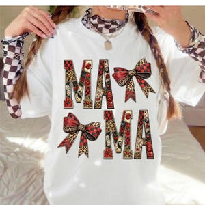 Può includere: T-shirt bianca con la scritta "MAMA" in un carattere decorativo. Le lettere sono decorate con motivi rossi e neri, ciliegie e labbra. Sono presenti anche due fiocchi con motivi leopardati e a scacchi.