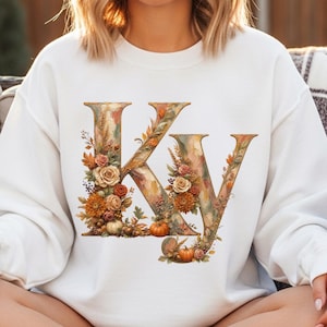Pode incluir: Camisola branca com as letras "KY" num design floral. As letras são adornadas com elementos outonais, incluindo abóboras, rosas e folhagem de outono em tons de laranja, castanho e verde. A estética geral é uma mistura de rústico e floral.