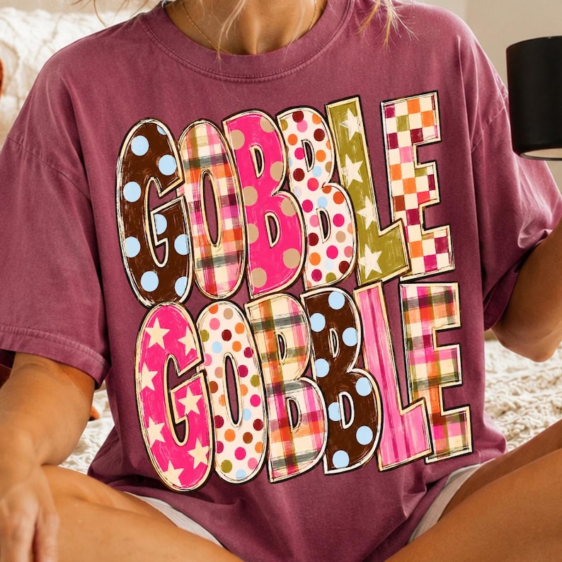 May include: A maroon short-sleeve t-shirt featuring the text GOBBLE GOBBLE printed in large, colorful letters. Each letter displays a different pattern, including polka dots, plaid, and stars in shades of pink, brown, orange, green, and blue.