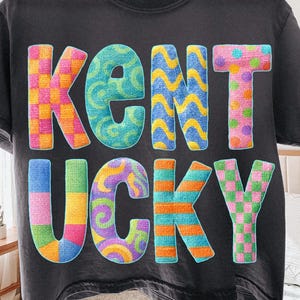 May include: Dark gray t-shirt with the word "KENTUCKY" in colorful, textured letters. The letters feature various patterns, including plaid, swirls, and stripes in shades of blue, pink, orange, and green.