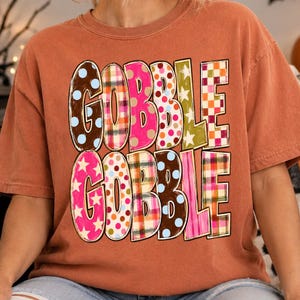 May include: A rust-colored short-sleeve t-shirt featuring the text GOBBLE GOBBLE printed in two rows. Each letter is filled with a different colorful pattern, including polka dots, plaid, and stars in shades of pink, brown, orange, and green.