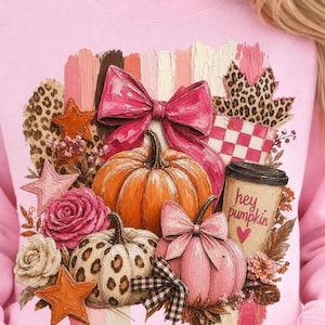 May include: Pink sweatshirt featuring a fall-themed graphic. The design includes pumpkins, bows, stars, and floral elements. The graphic also has a coffee cup with the text "hey pumpkin" and a heart.