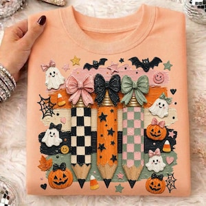K&ouml;nnte beinhalten: Pfirsichfarbenes Sweatshirt mit einem Halloween-Motiv. Das Design umfasst drei bleistiftf&ouml;rmige Dekorationen mit Schleifen, Flederm&auml;usen, Geistern, K&uuml;rbissen, Sternen und Spinnweben. Die Bleistifte haben karierte und Sternmuster.