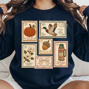 May include: Navy blue sweatshirt featuring vintage-style postage stamp designs. Stamps depict a pumpkin, duck, acorn, magnolia, bow, and a thermos. Text includes "Autumn Harvest," "Wild and Free," "Nature's Treasure," "Southern Bloom," "Sweet Southern Charm," and "Warm Days Cool Nights."